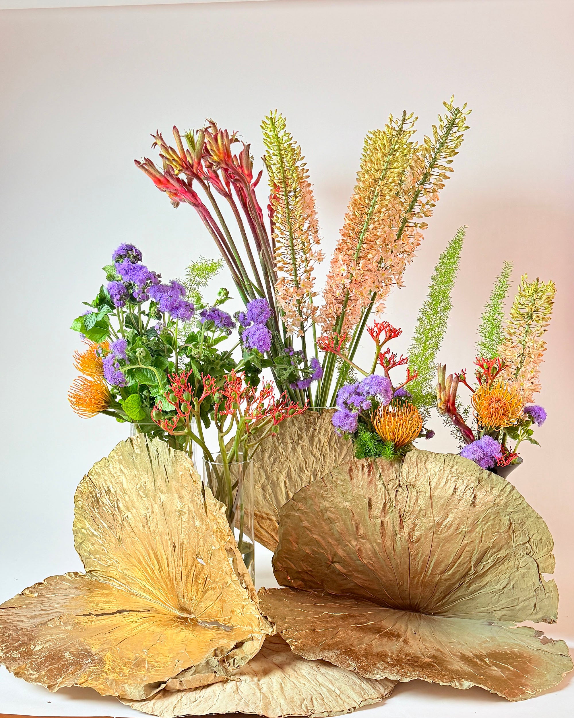 Composizione con fiori arancioni, viola e color pesca, verde ornamentale e grandi foglie dorate su fondo chiaro.