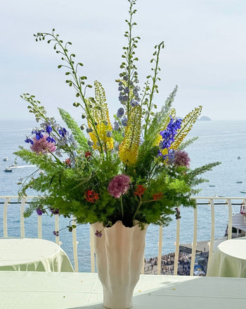 Composizione floreale con fiori gialli, blu e viola e verde ornamentale in vaso bianco su una terrazza vista mare